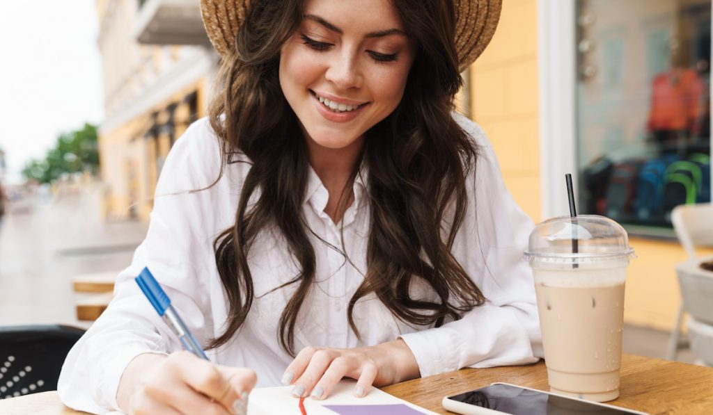 Portrait of smiling woman writing down in her notebook while drinking milkshake