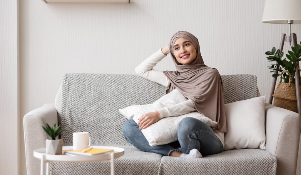 Relaxed arabic girl daydreaming in hijab sitting on sofa