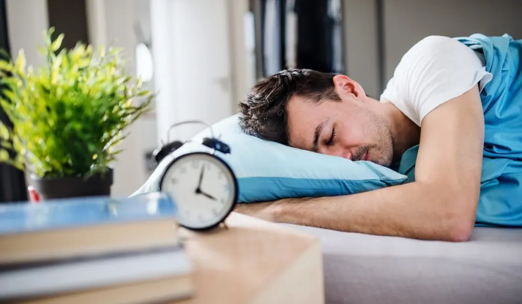 Young man with alarm clock in bed at home, sleeping