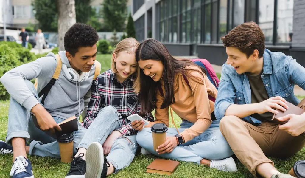 happy teenagers sitting on grass looking at phone in the school