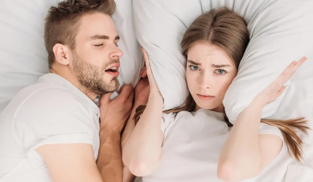 top view of woman plugging ears with pillow while lying in bed near snoring husband