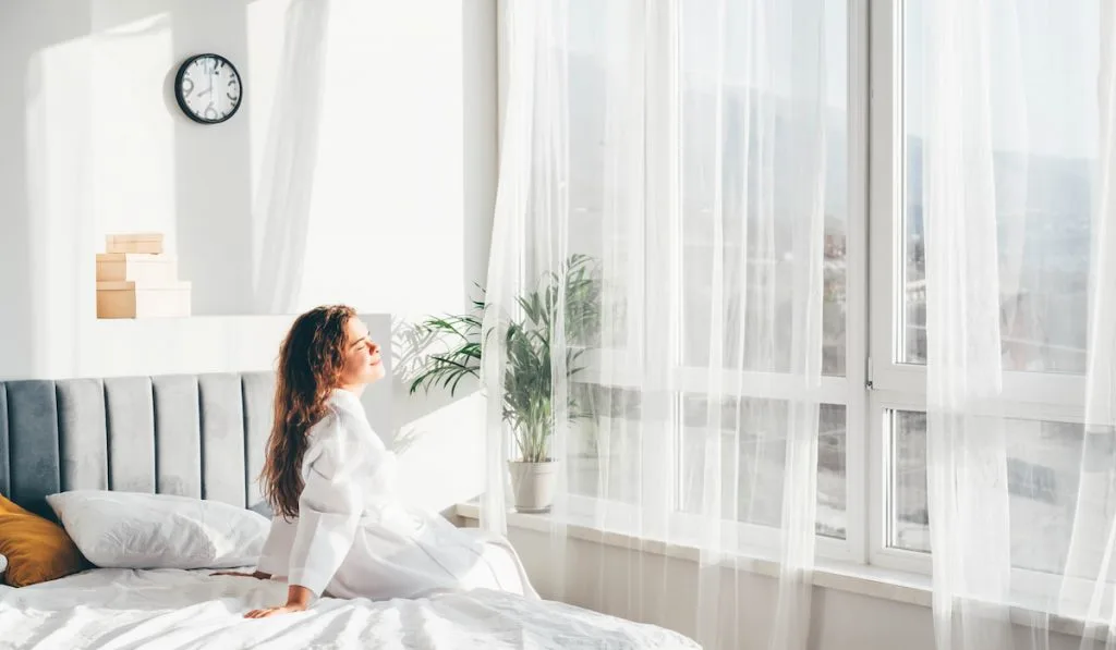 woman in bathrobe sitting near the big window on bed after waking up 