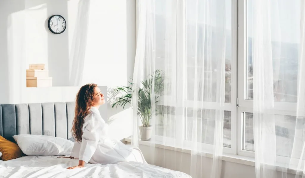 woman in bathrobe sitting near the big window on bed after waking up